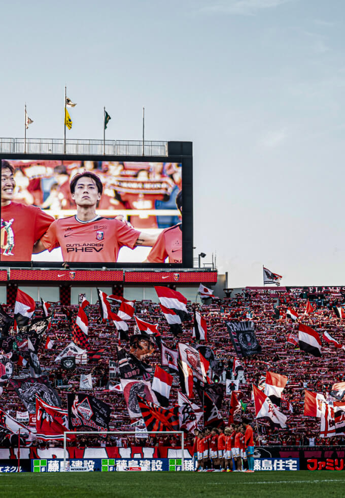 試合開始直前、ピッチには選手たちが一列に並び、その後ろに見えるゴール裏のスタンドには旗を振る多くの観客たちが見える。