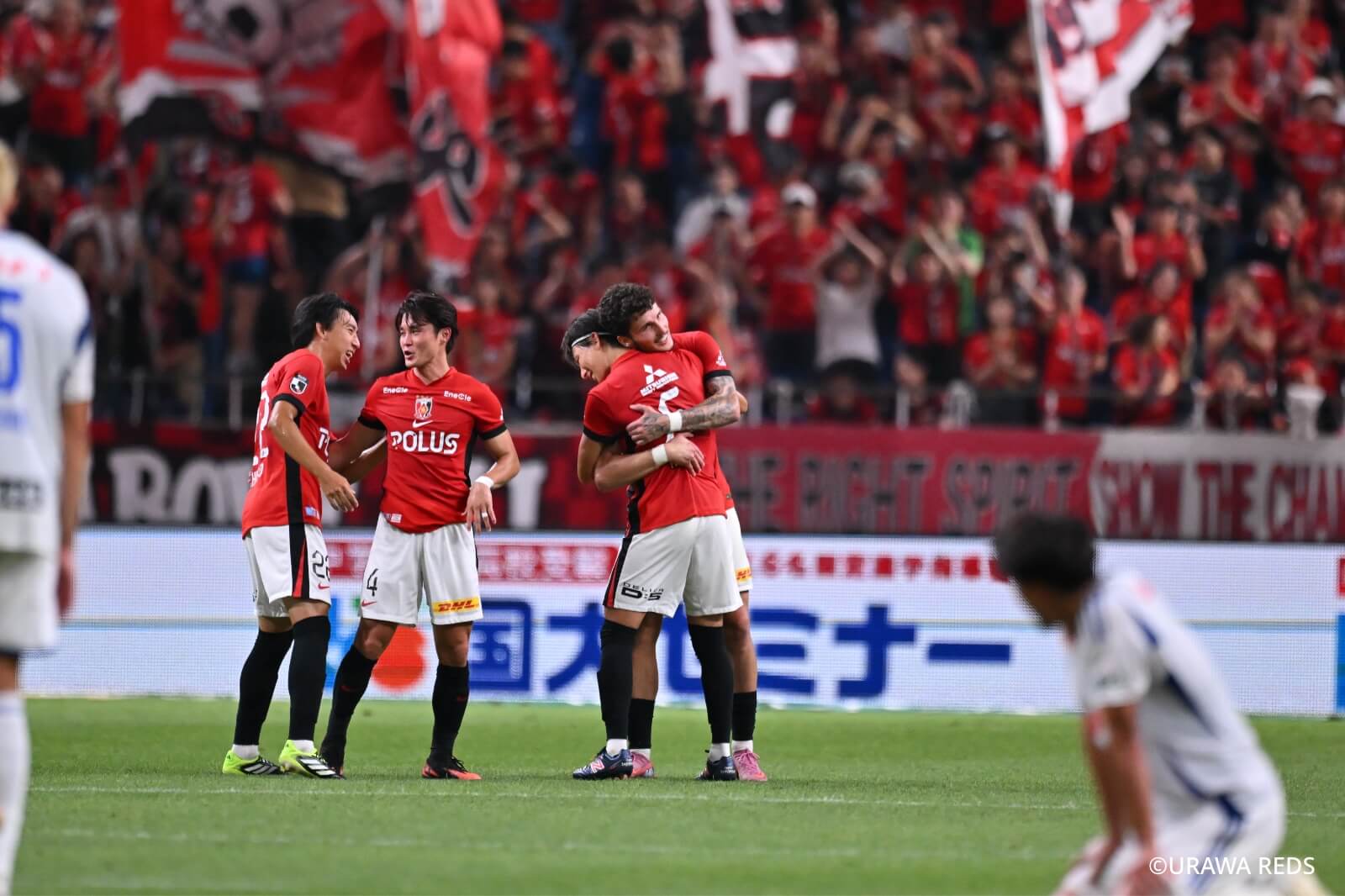 試合中、ピッチで抱擁を交わす浦和レッズ選手たちと、それを膝をつき遠目で眺める相手選手